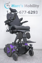 A black power wheelchair with purple accents, photographed indoors against a white wall with a Marc's Mobility sign in the background.