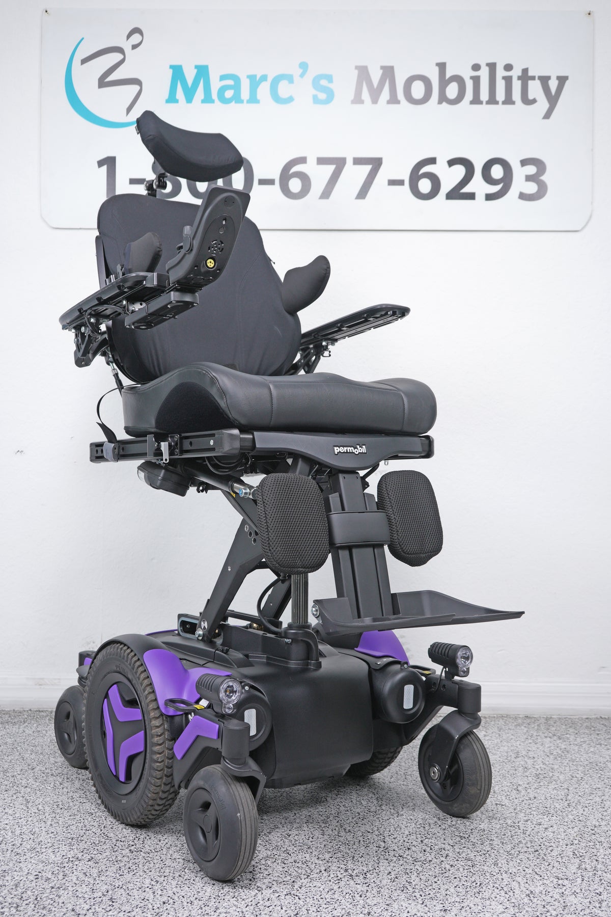 A black power wheelchair with purple accents, photographed indoors against a white wall with a Marc's Mobility sign in the background.