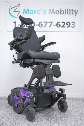 A black power wheelchair with purple accents, photographed indoors against a white wall with a Marc's Mobility sign in the background.